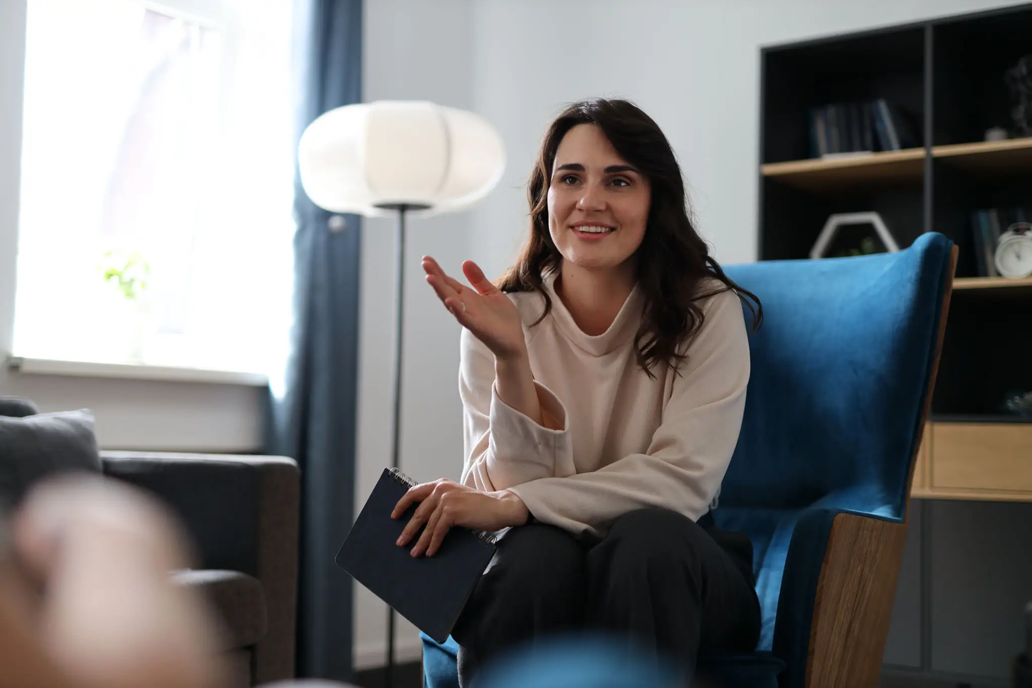 Woman sitting on a blue chair, smiling and gesturing with her hand.