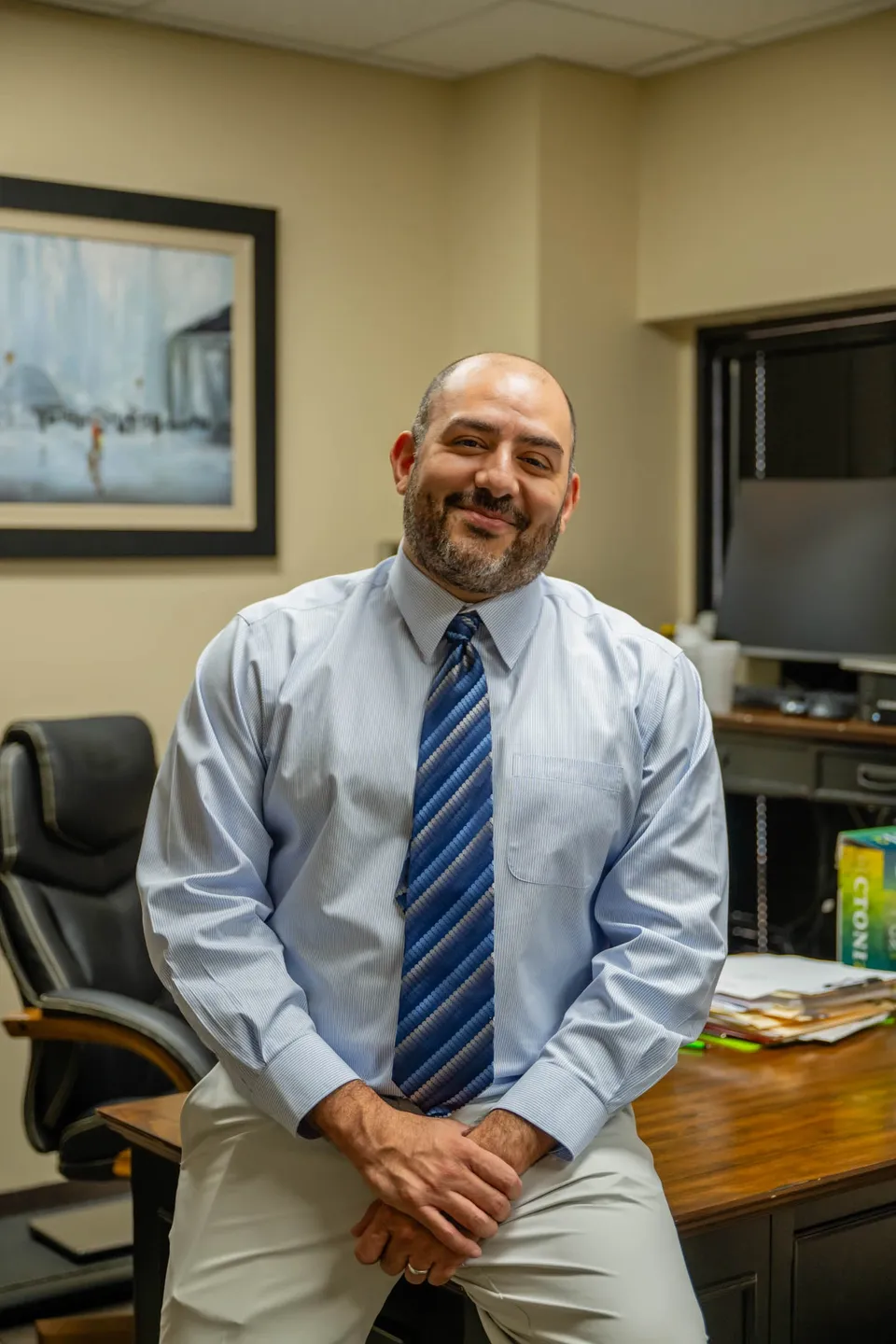 Man in office attire smiling while sitting on a desk.